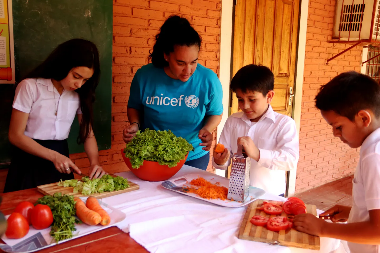 Ni&ntilde;os y ni&ntilde;as preparando una ensalada, cortando tomates, lechuga y zanahorias, con la ayuda de una trabajadora de UNICEF 