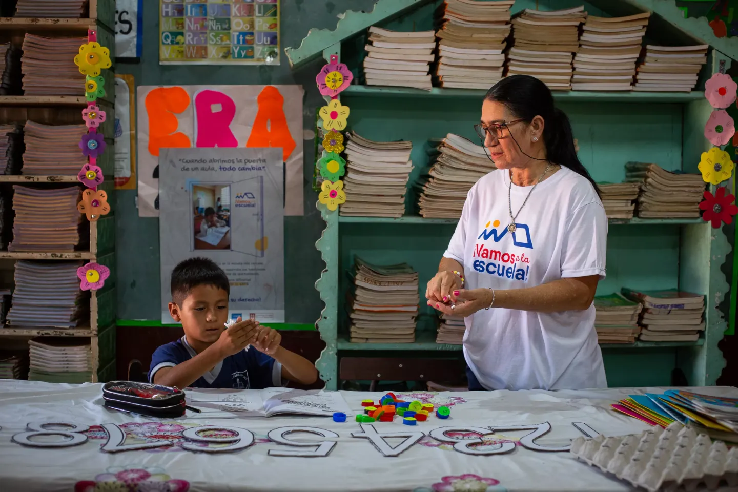 Ni&ntilde;o aprendiendo con materiales did&aacute;cticos junto a su maestra en un aula decorada y colorida.