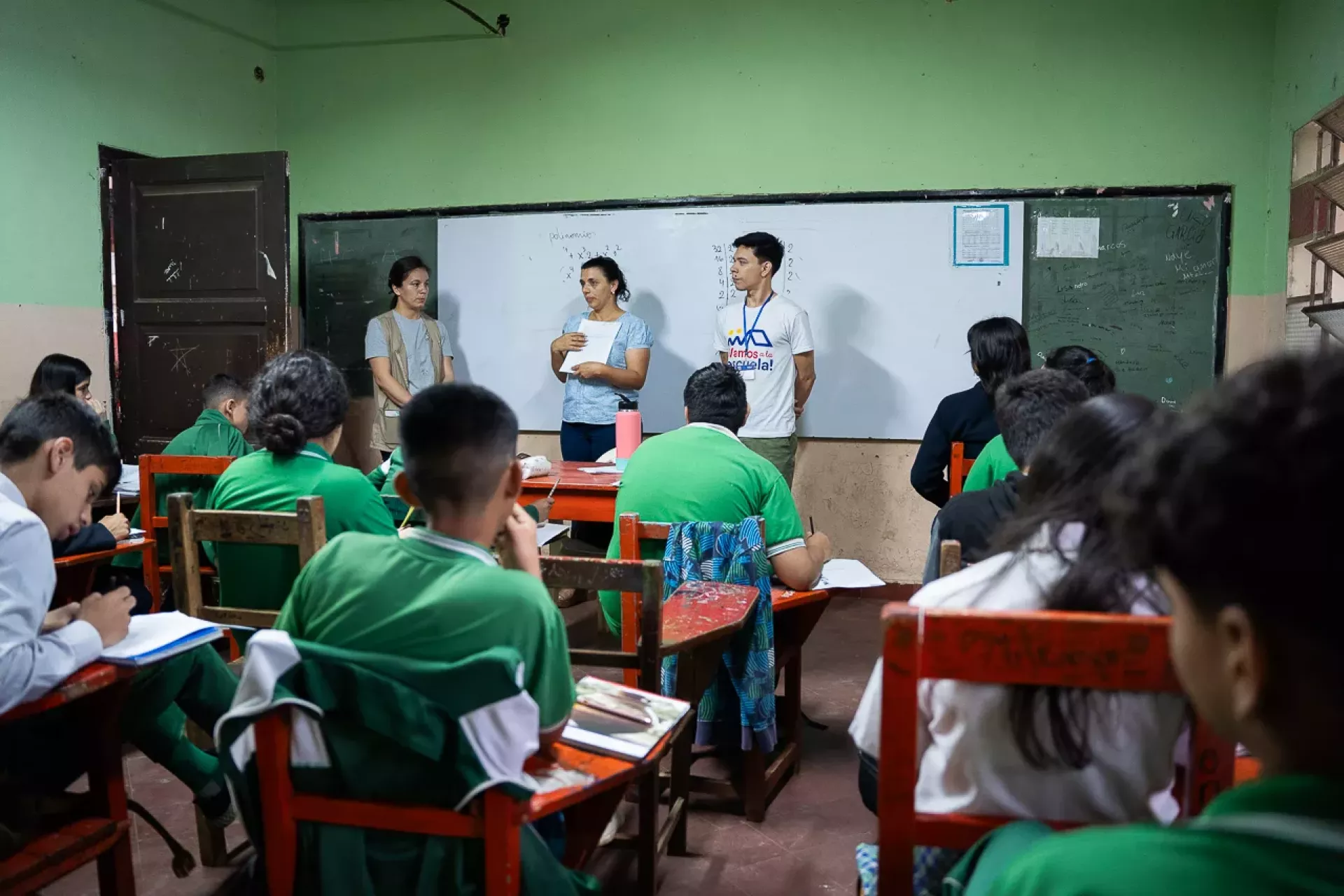 En una sala de clases, dos voluntarios y una maestran se paran frente al pizarr&oacute;n para explicar sobre la evaluaci&oacute;n