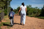Mujer y ni&ntilde;o caminando de la mano por un camino de tierra, el ni&ntilde;o lleva una mochila escolar azul.