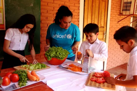 Niños y niñas preparando una ensalada, cortando tomates, lechuga y zanahorias, con la ayuda de una trabajadora de UNICEF 