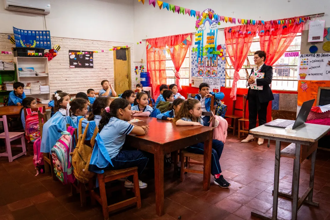 Grupo de estudiantes participa en una actividad educativa en el aula, guiada por una docente y apoyada por herramientas digitales para el aprendizaje.