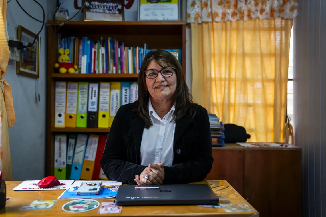 Persona sentada en un escritorio dentro de una oficina escolar, con una computadora port&aacute;til frente a una estanter&iacute;a con libros y carpetas.