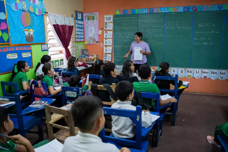 En un sal&oacute;n de clases los estudiantes est&aacute;n sentados atendiendo a la maestra que est&aacute; parada al frente explicando 