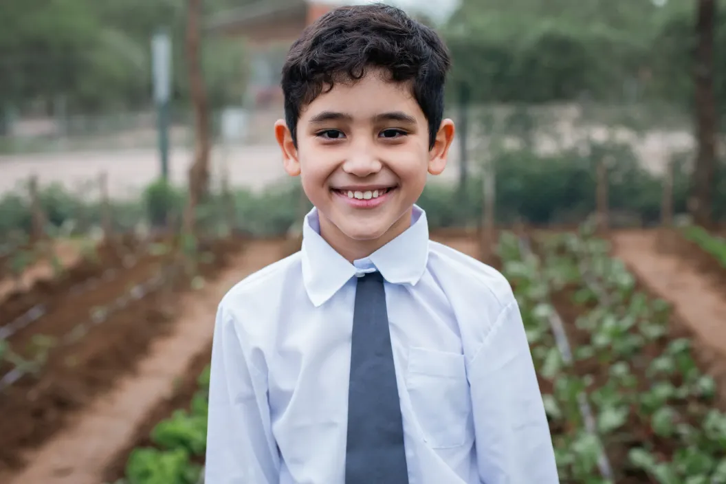 Ni&ntilde;o con camisa blanca y corbata gris sonriendo, detras se ve la huerta escolar