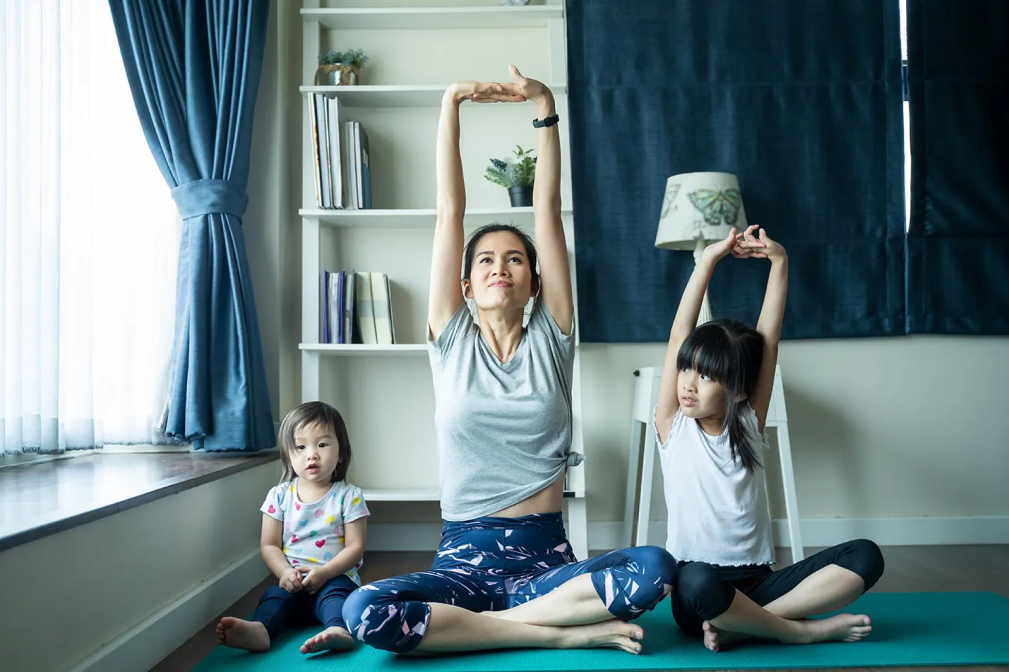 Mother and an infant and older girl sitting next to her on the floor stretching their arms.