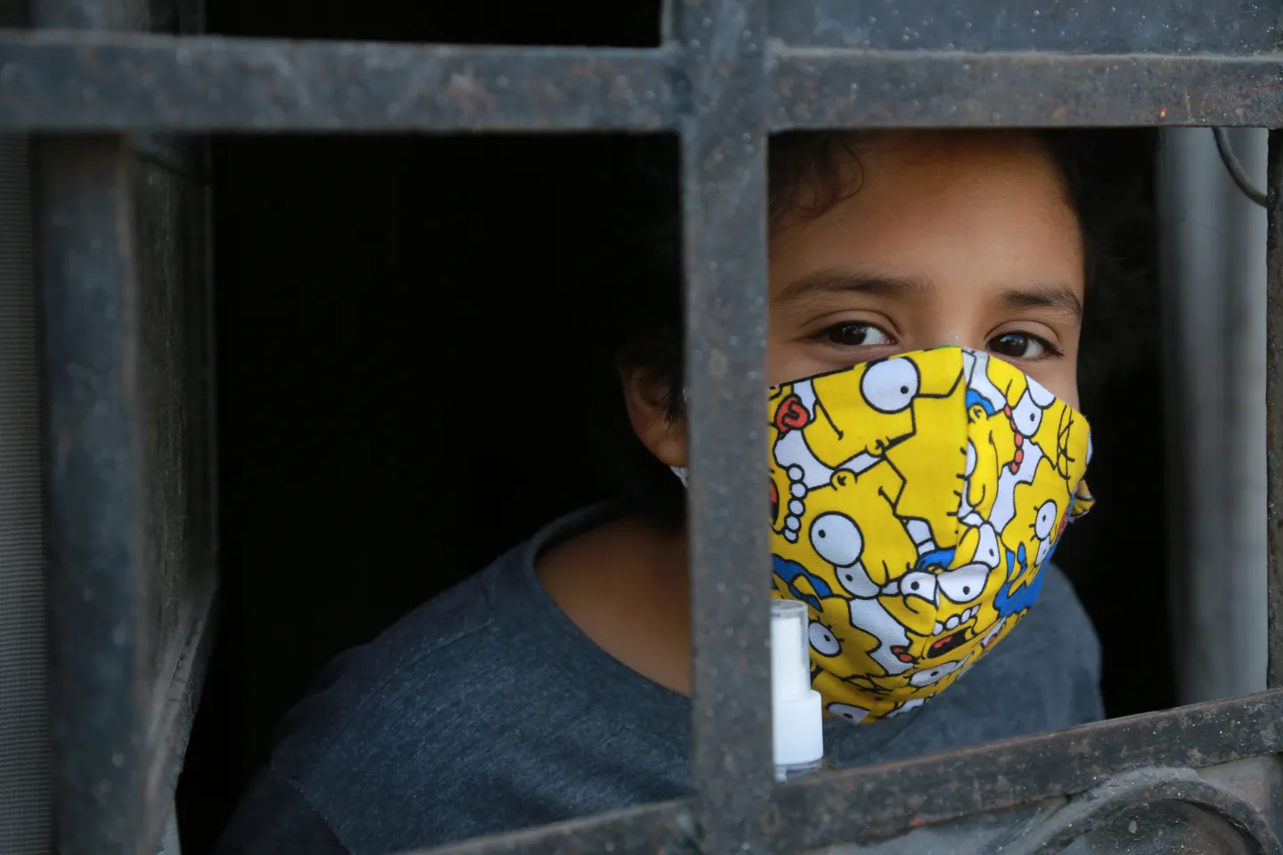 Un nino usando mascarilla, mira a traves de su puerta hacia la calle