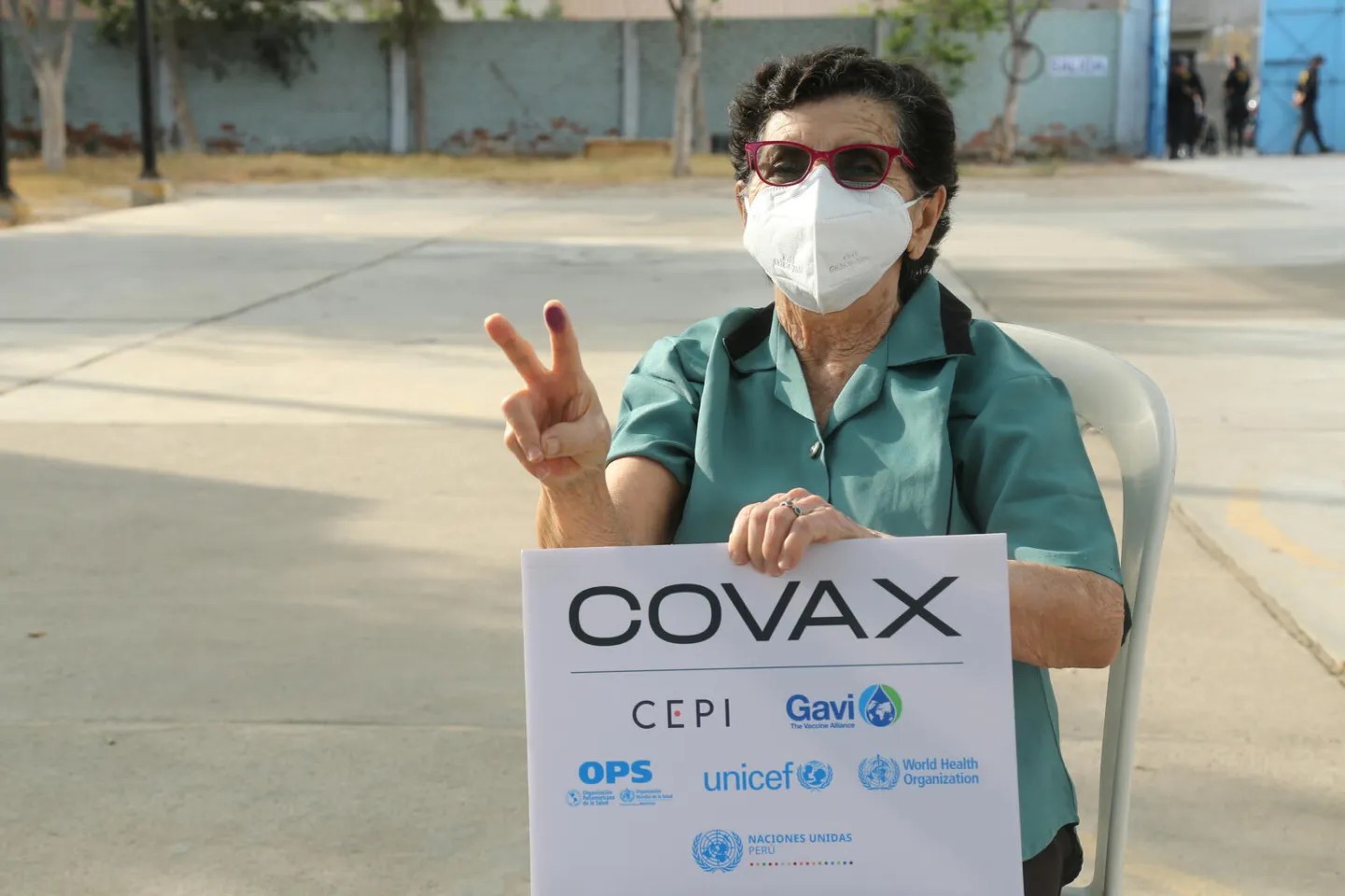Una mujer, adulta mayor, hace el s&iacute;mbolo de V, en referencia a vacunas, mientras levanta un cartel de COVAX