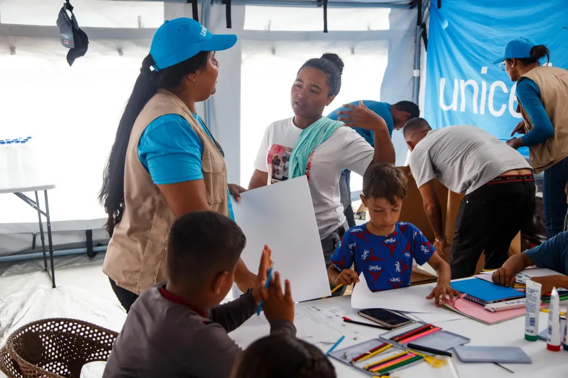 Un adolescente habla con una mujer adulta, dentro de un carpa donde otros ninos realizan actividades como dibujo y pintura.
