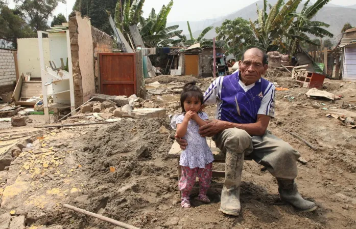 Un adulto mayor y una nina sentados frente a una casa destruida por un aluvi&oacute;n