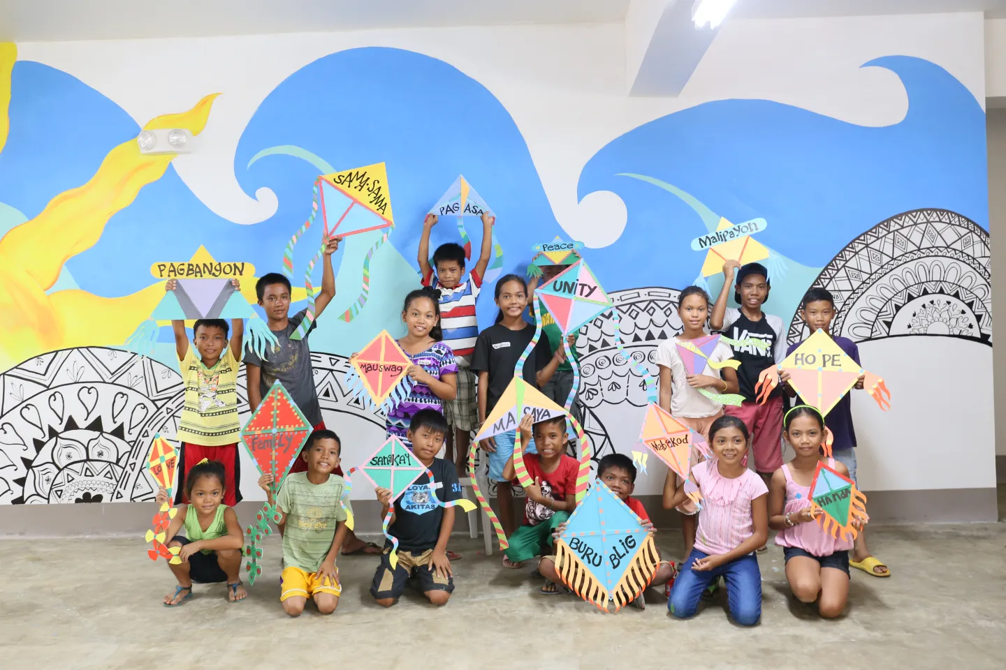 Children hold paper kites with inspirational words while standing in front of a mural that they painted