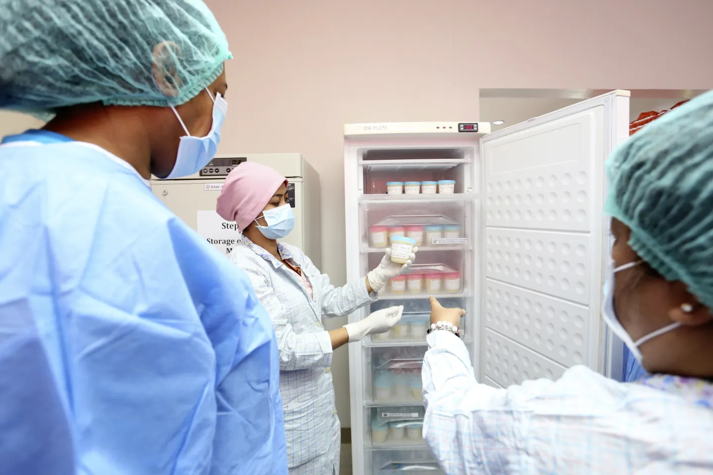 Health workers wearing laboratory gowns, gloves, hair caps and face masks inspect breast milk stored in a refrigerator at a human milk bank
