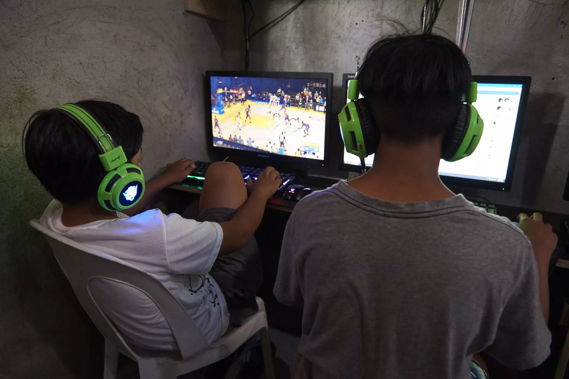 Two children playing games inside a public computer rental shop. Their faces are turned away from the camera, hiding their identities