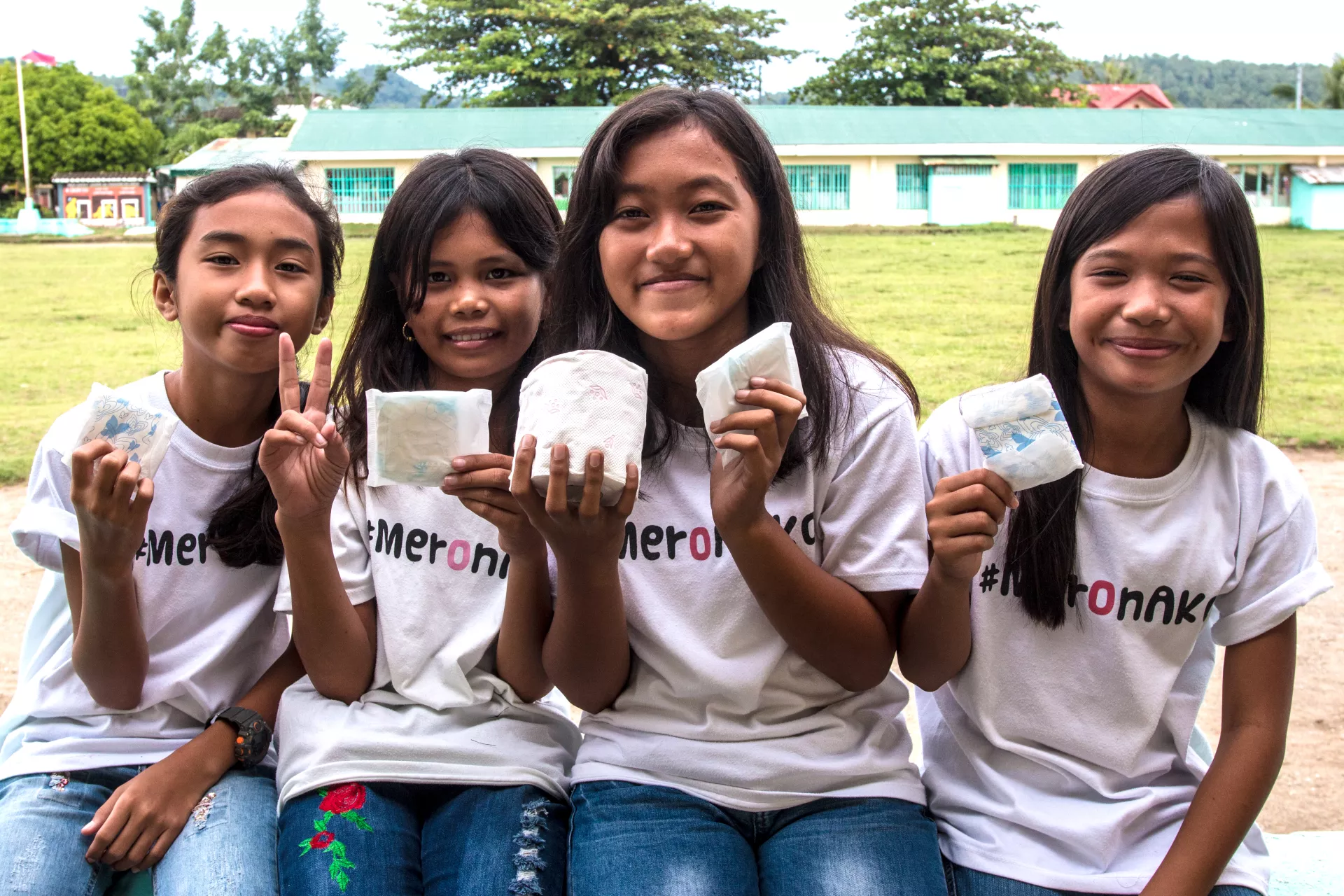 Four girls, wearing t-shirts with the hashtag #MeronAko, holding sanitary napkins