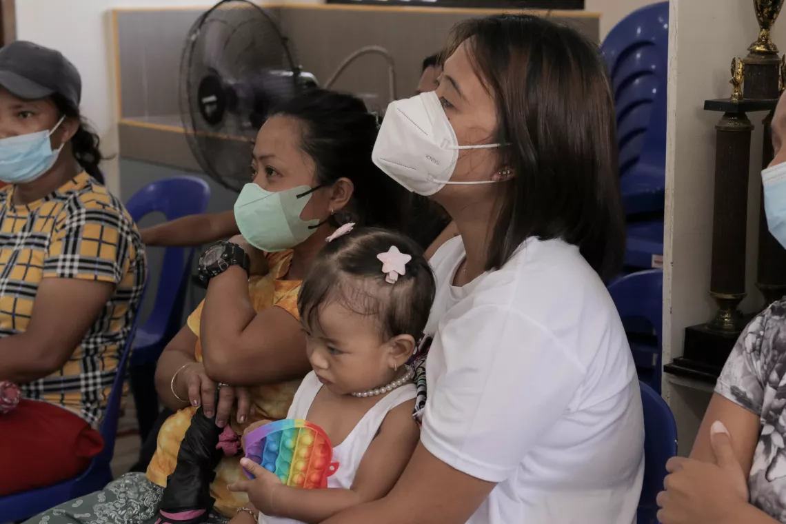 A mother listens to an information session on infant and young child feeding as she holds her daughter on her lap. The mother and another adult visible in the photo are wearing face masks.