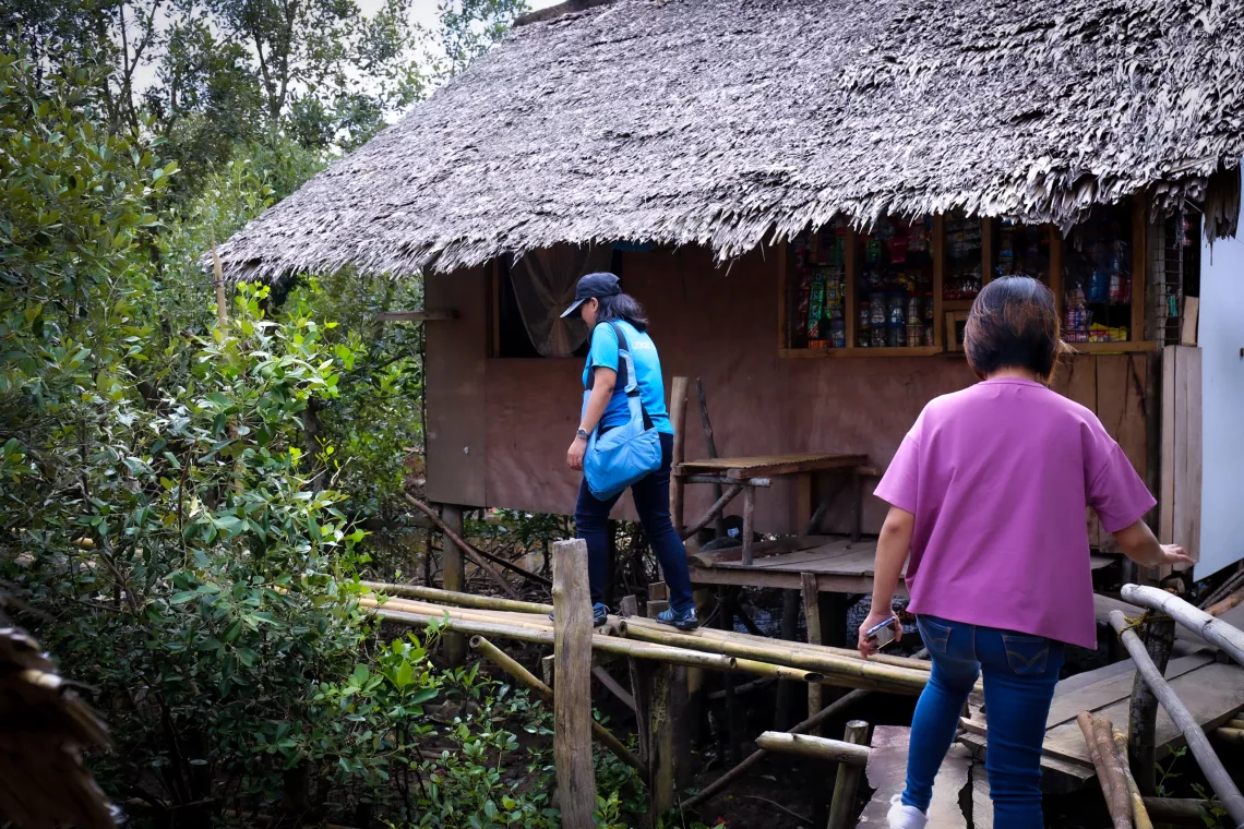 A UNICEF staff member in a cyan t-shirt and a health worker crossing a narrow bridge made of bamboo poles