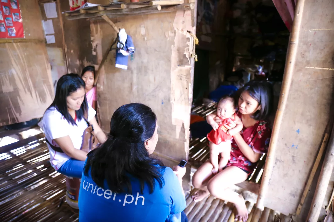 A UNICEF staff member in a blue UNICEF t-shirt and a health worker speaking with a young mother holding her baby at her home