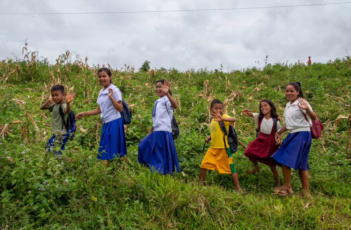 Children carrying backpacks and wearing school uniforms walking across a grassy field and waving to the camera