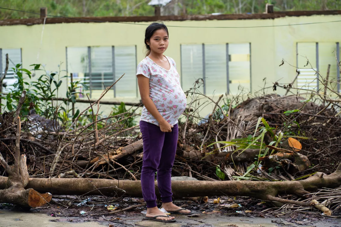 A pregnant adolescent standing amid debris in front of a damaged building