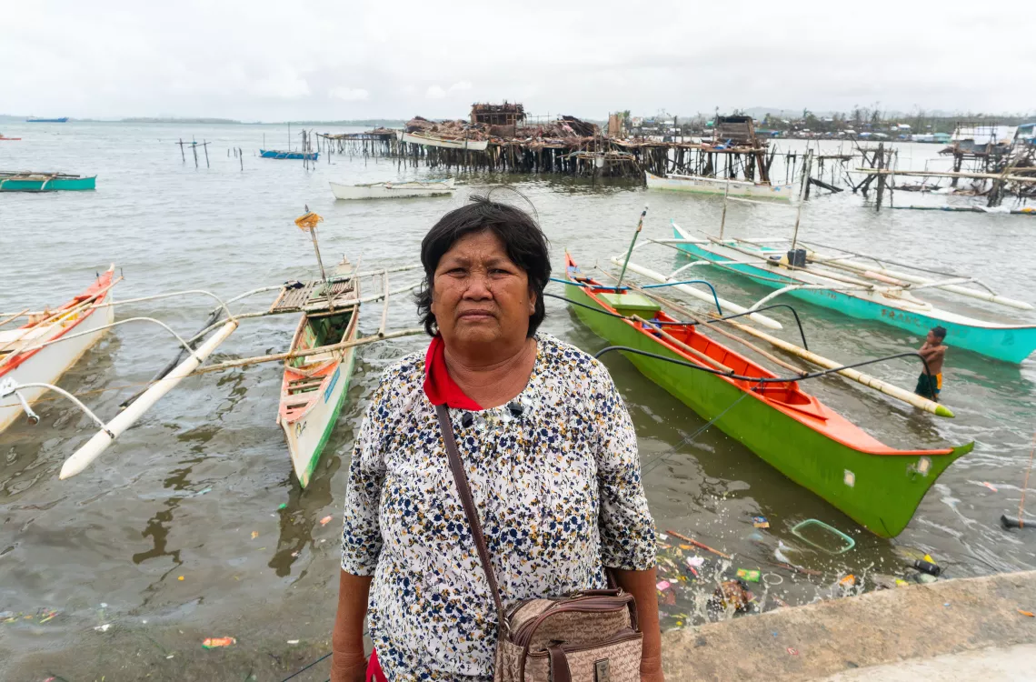 A woman standing by the port shore, with fishing boats and stilt houses in the background