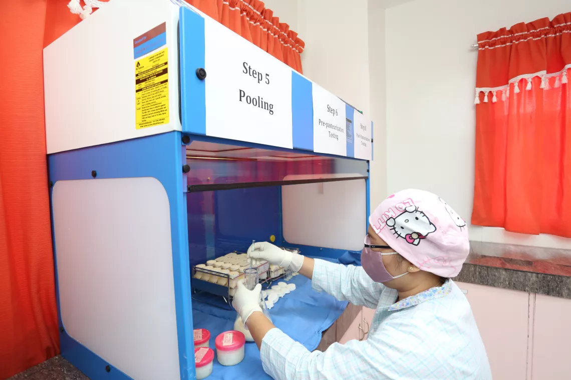 A health worker, wearing a laboratory gown, gloves, face mask and hair cap, transfers breast milk to a new container under a laminar flow hood.