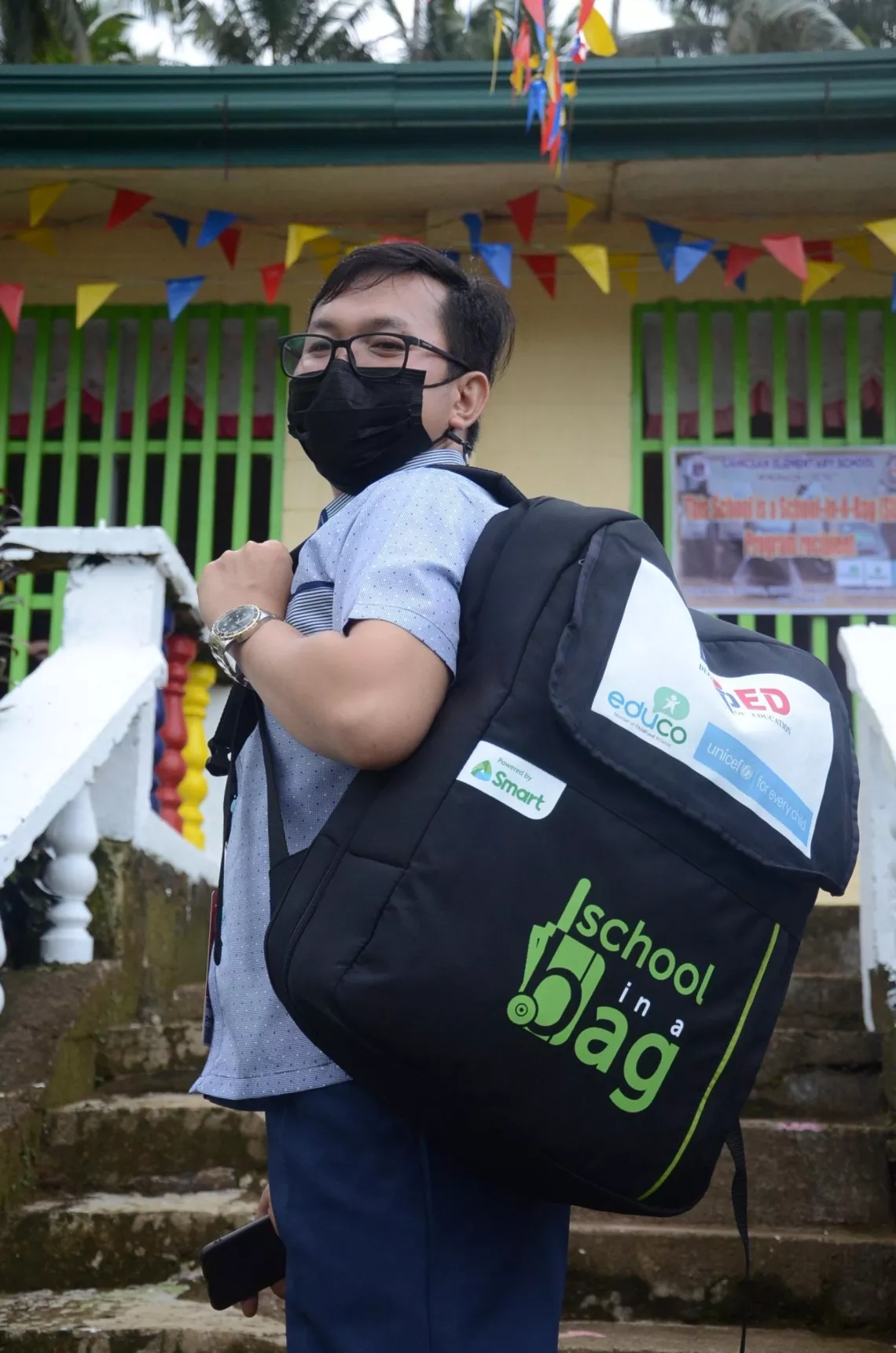 A teacher carrying a backpack labeled 'School-in-a-Bag'