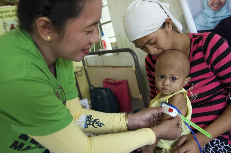 A health worker measures the circumference of a child's upper arm using a mid-upper arm circumference (MUAC) tape to determine the child's nutrition status. The pointer of the tape is in the red figures range, which means that the child is thin for their height.