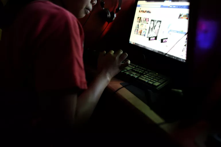 A girl, her face hidden, browses Facebook inside a dark internet cafe