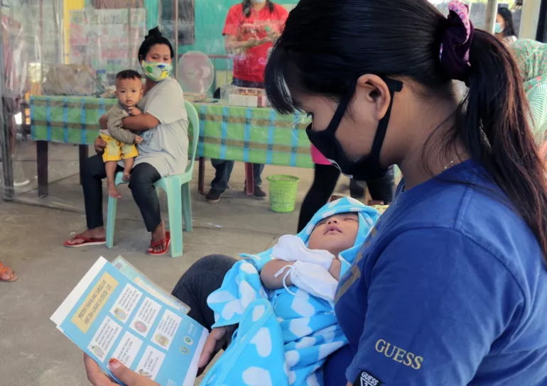 A mother wearing a mask is reading a leaflet while carrying her baby