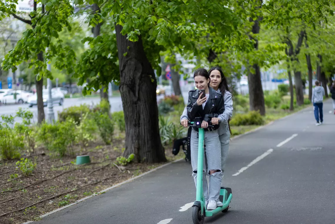 Două adolescente merg&acirc;nd &icirc;mpreună pe o trotinetă electrică. 
