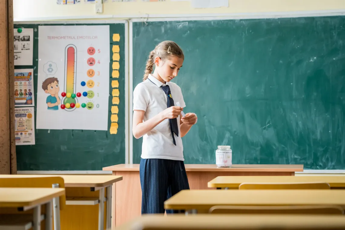 Elevă &icirc;ntr-o sală de clasă care citește un bilețel &icirc;n fața tablei. 