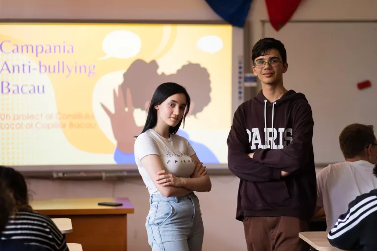 A boy and a girl pose in front of their presentation at a workshop they are delivering for peers.