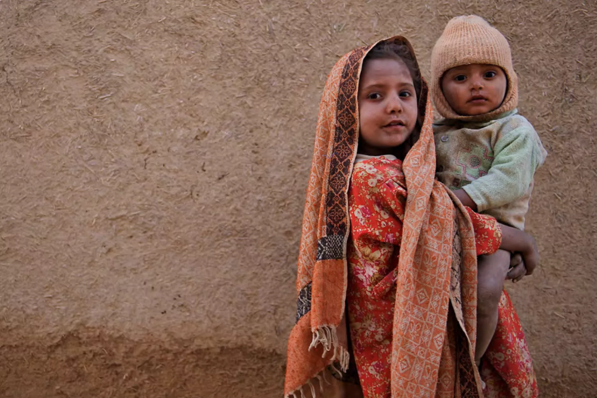 A girl holds a small child with a plaster wall in the background