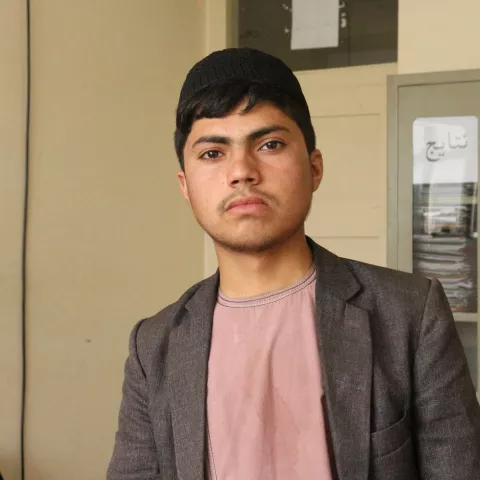 Naqibullah, an adolescent boy, looks at the camera with determination. He is wearing a black pakol, light red shirt and grey-brown sports jacket. A beige wall is in the background.