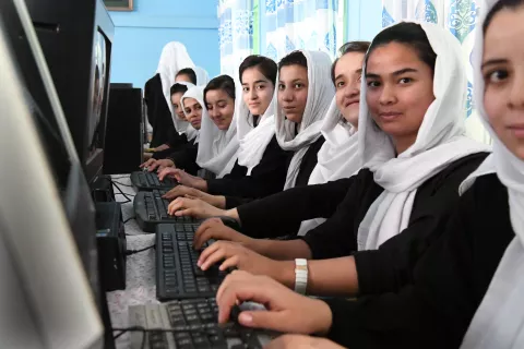 A like of girls in their black school uniforms and white hijabs work on computers while they look at the camera