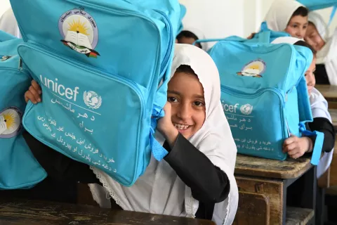 A young girl in a white hijab sits behind a desk and holds up a bright-blue UNICEF backpack, smiling
