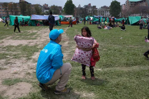 A girl stands holding a pillow talking to a UNICEF worker, field of tents in the background, 