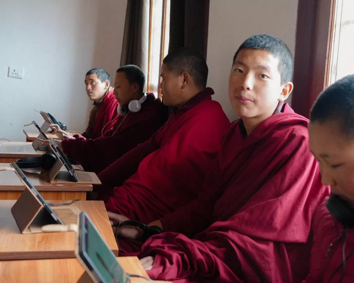 An adolescent student monk looks up at the camera as his fellow students work on their digital learning tablets.