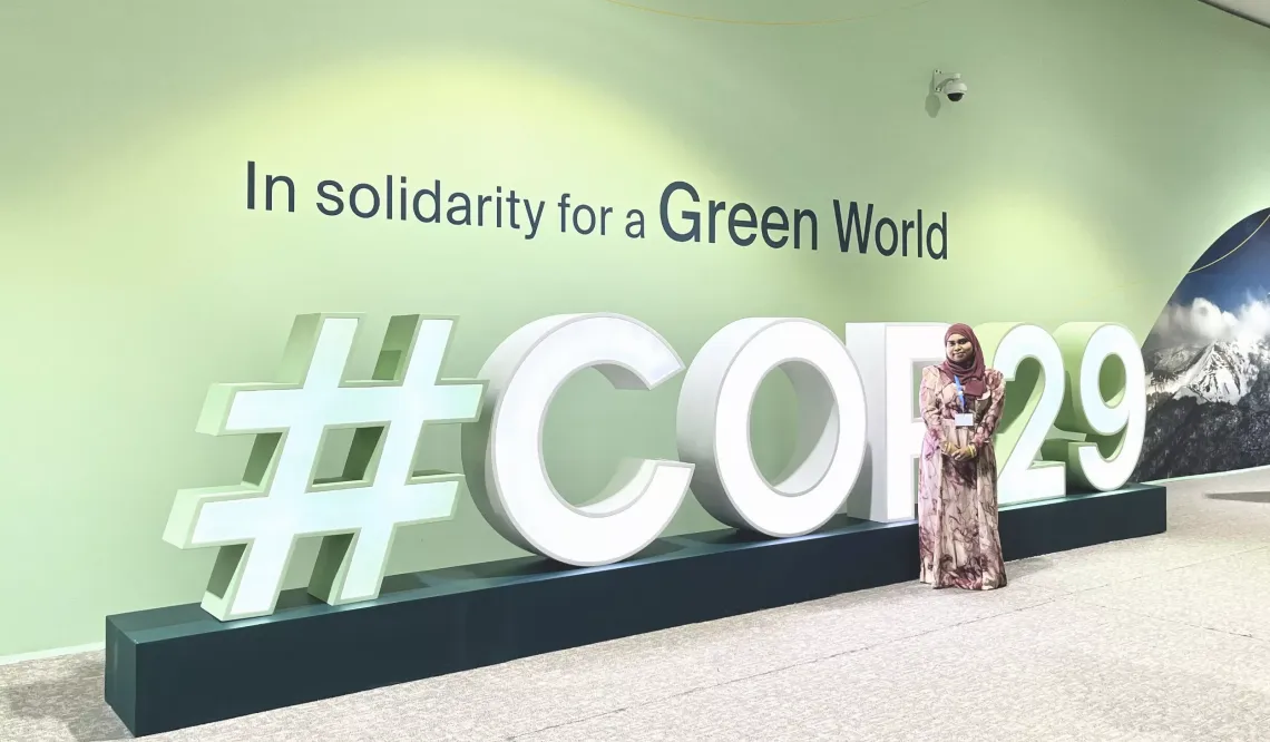 Fathima, UNICEF Maldives Youth Advocate standing in front of a green wall with the words: "In Solidarity for a Green World COP29"