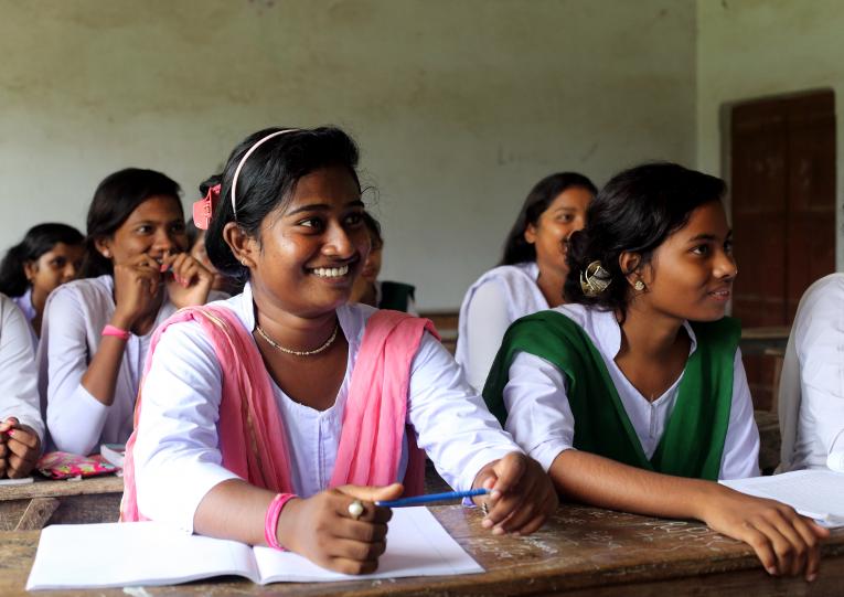 Adolescent girls smile away from the camera in a classroom, their books lay on their desks.