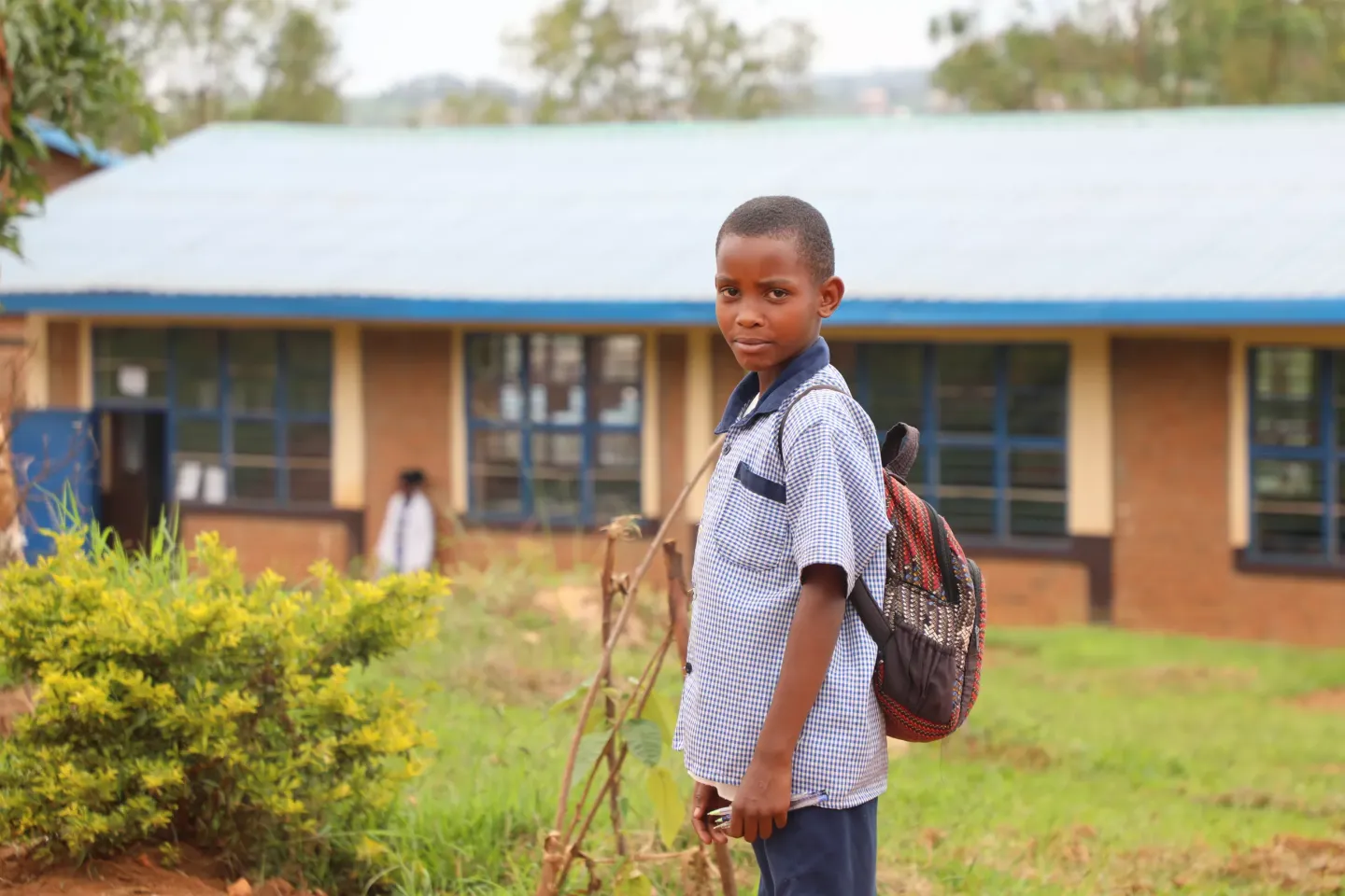 A boy in blue school uniform looks to the camera with a classroom block in the background. 