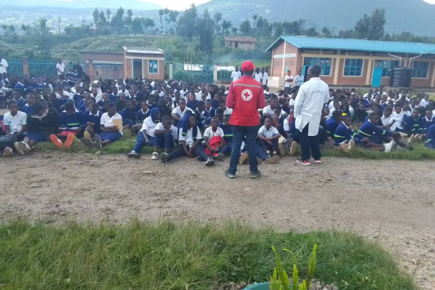 A man dressed in a red shirt with the Red Cross insignia on it, addressing a crowd of people who are seated. 