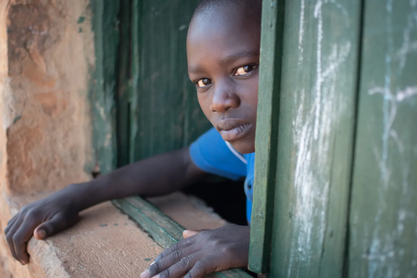 A young boy in a school uniform peeks out of a wooden schoolhouse window