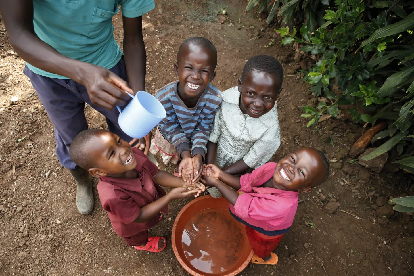 Several small children stand around a bucket of water while an adult male pours water from a cup so they can wash their hands.
