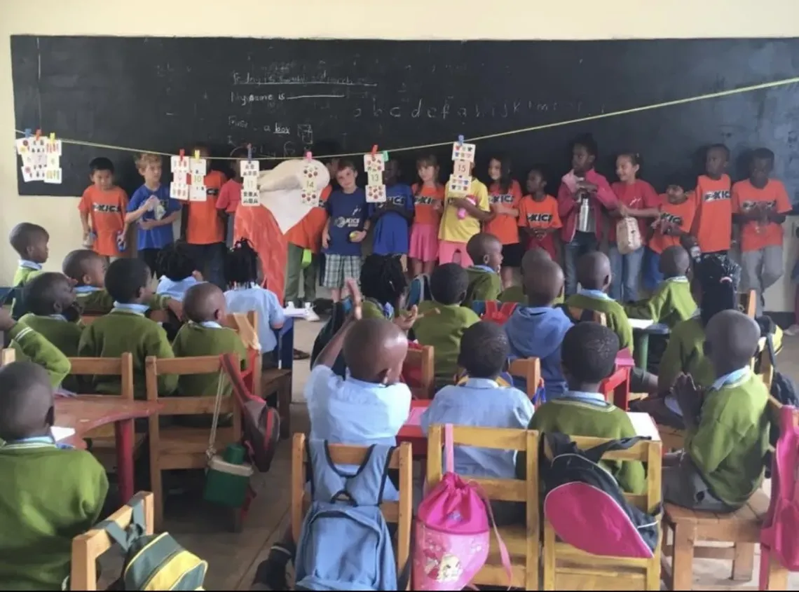 Children watch classmates perform on a classroom stage in front of a chalkboard, colorful backpacks on chairs.