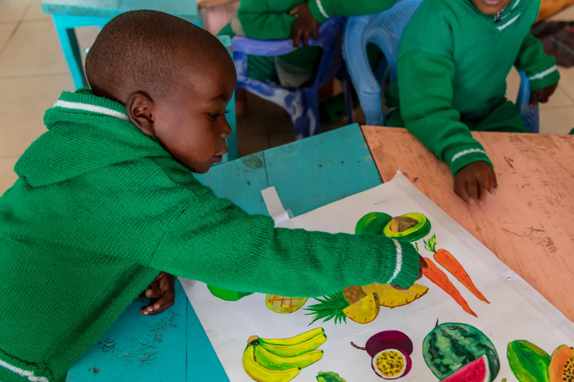 David, 6, points to a painting of a carrot on a poster showing different fruits and vegetables. He sits at a small blue table inside his classroom at the Rutsiro Tea Plantation day care centre, supported by UNICEF.