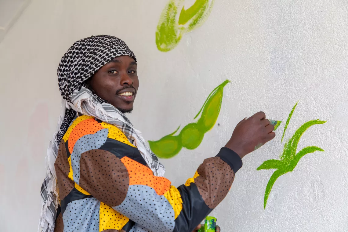 Timothy Wandulu, painter and sculptor, smiles into the camera while posing with a paintbrush. He is working on a mural depicting fruits and vegetables, currently painting green on a picture of a bean pod.