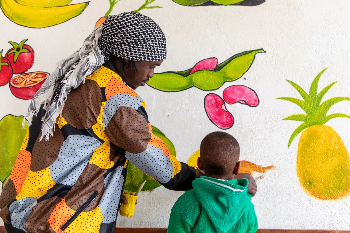 Timothy Wandulu, painter and sculptor, helps a little boy at the UNICEF supported Rutsiro early childhood development center to paint fruit on a mural. les, currently painting green on a picture of a bean pod.