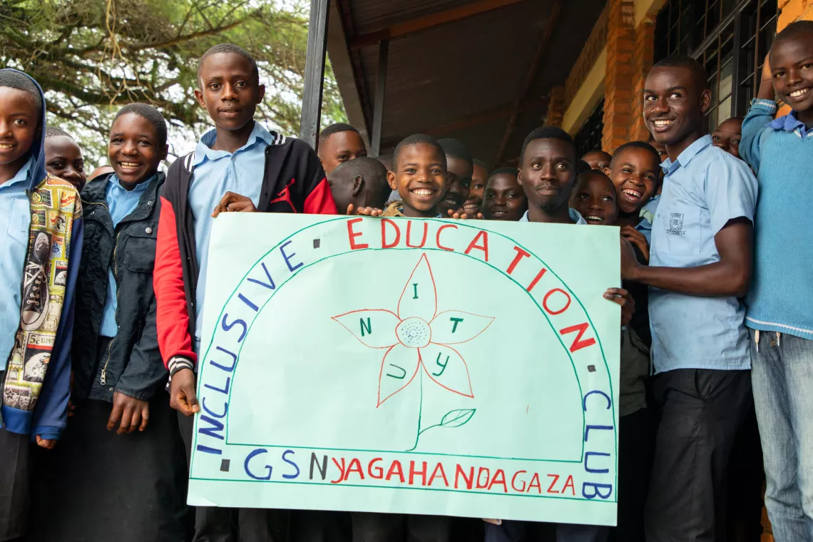 A group of students hold a hand-drawn sign that says "Inclusive Education Club" and smile. A few days a week, students with and without disabilities meet for Inclusive Education Club to play games together, dance and sing.