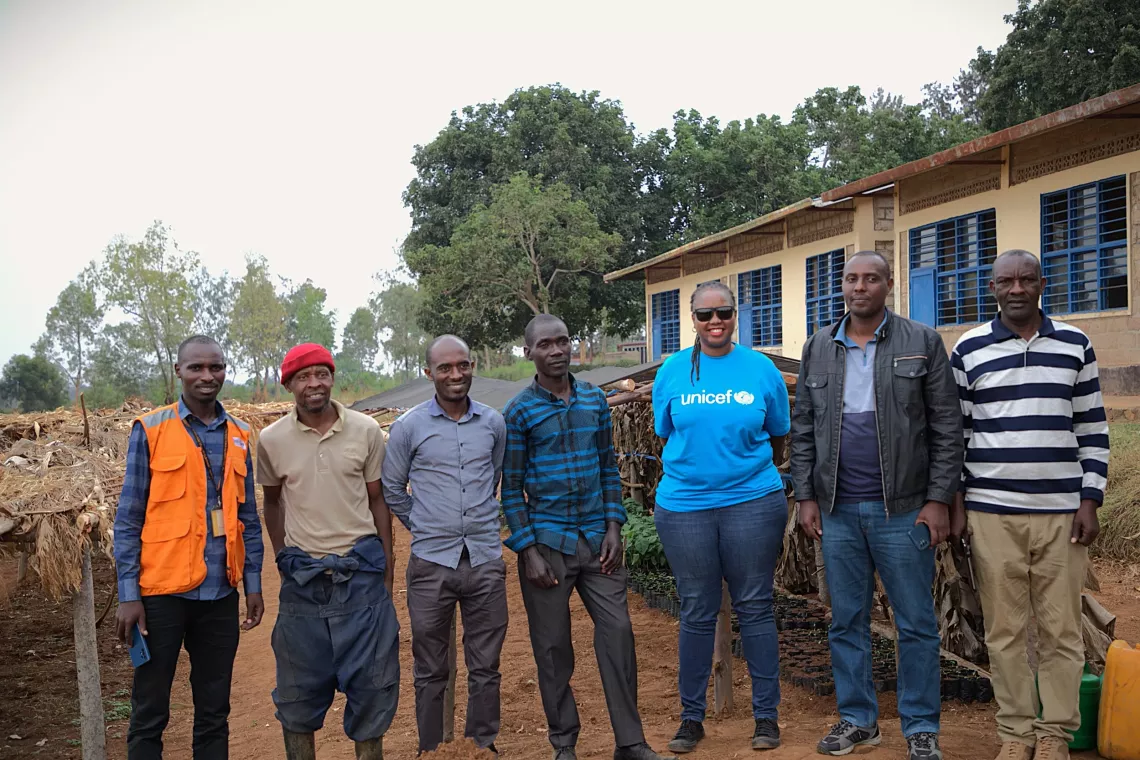 Six men and one woman gather in a line for a picture with a seedling nursery behind them.